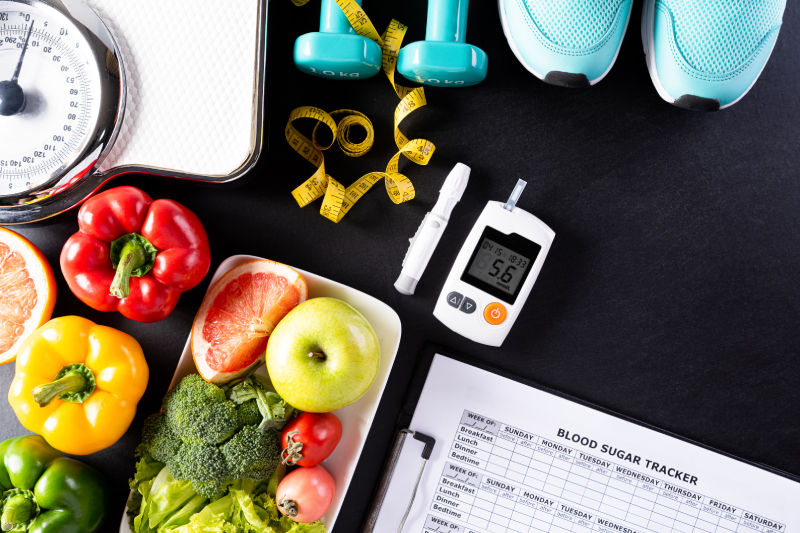 An arrangement of fruits, vegetables, a glucometer, dumbbells, shoes, a scale, and a sugar tracker sheet representing a diabetes care routine.