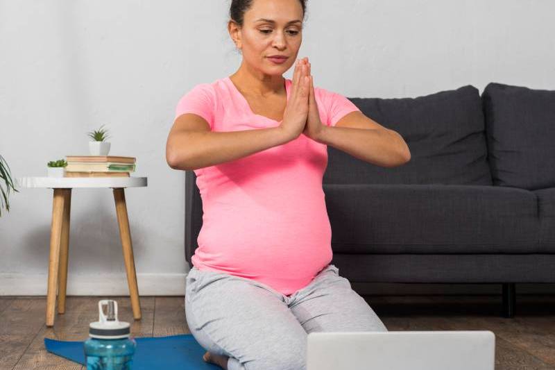 A pregnant woman practicing online yoga at home using a laptop.