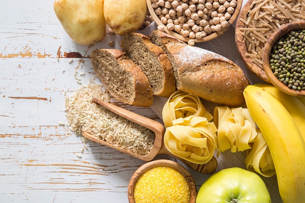 Complex Carbohydrate-rich foods including bread, rice, pasta, bananas, potatoes, pulses, and fruits on a table.