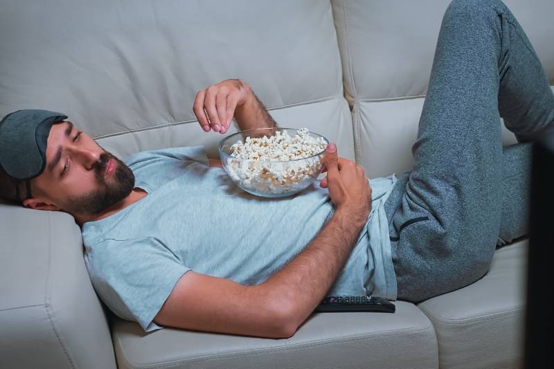 Man on sofa eating popcorn and watching TV, illustrating sedentary habits linked to diabetes.
