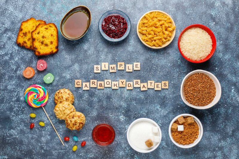 Simple Carbohydrate foods such as pasta, rice, cookies, syrup, candies, and sugar cubes arranged on a table.