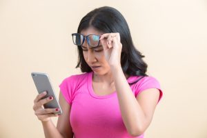 Woman lifting her glasses and straining to read her phone, suggesting a need for anti-reflective lenses.