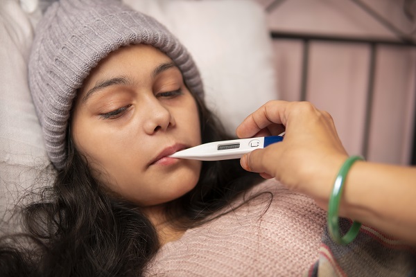 A girl wearing a sweater and cap checks a thermometer, showing symptoms of fever.