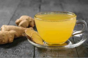 A cup of yellow ginger tea with several slices on a saucer, placed next to fresh ginger root.