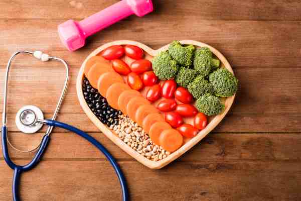 Heart-shaped plate with vegetables, stethoscope, and dumbbell, representing healthy eating, exercise, and stress control for heart health.