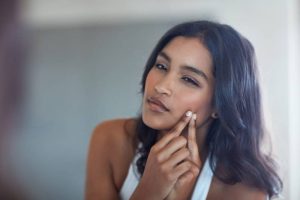 A woman pinching a pimple on her cheek in the mirror, symbolizing breakouts caused by hormonal changes.