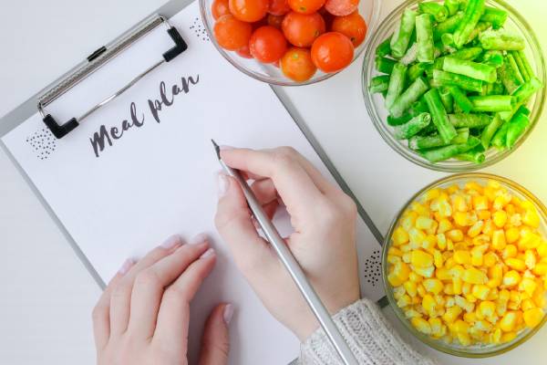 Person writing a meal plan with fresh veggies nearby, highlighting the importance of careful planning.